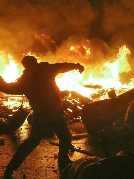 Vor einer brennenden Barrikade ist der Schattenriss eines Mannes zu sehen, der einen Gegenstand wirft. Vor einer brennenden Barrikade ist der Schattenriss eines Mannes zu sehen, der einen Gegenstand wirft.