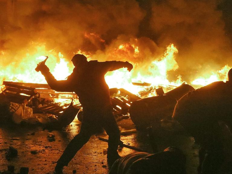 Vor einer brennenden Barrikade ist der Schattenriss eines Mannes zu sehen, der einen Gegenstand wirft. Vor einer brennenden Barrikade ist der Schattenriss eines Mannes zu sehen, der einen Gegenstand wirft.