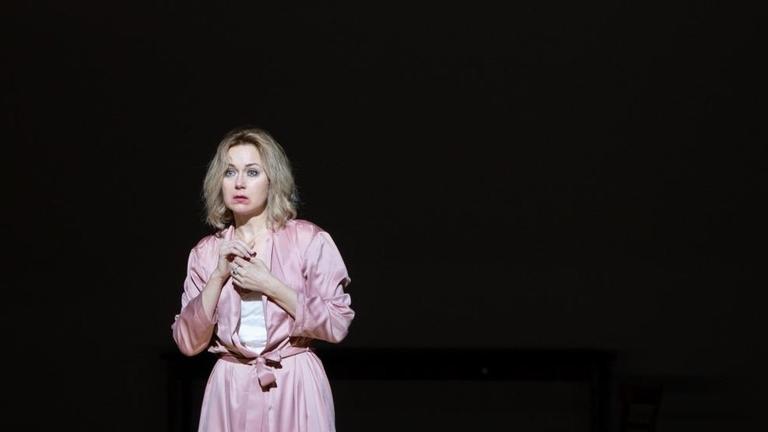 Eine junge Frau mit blonden Haaren steht in einem rosafarbenem Kostüm allein auf einer Bühne, die um sie herum stockdunkel ist.