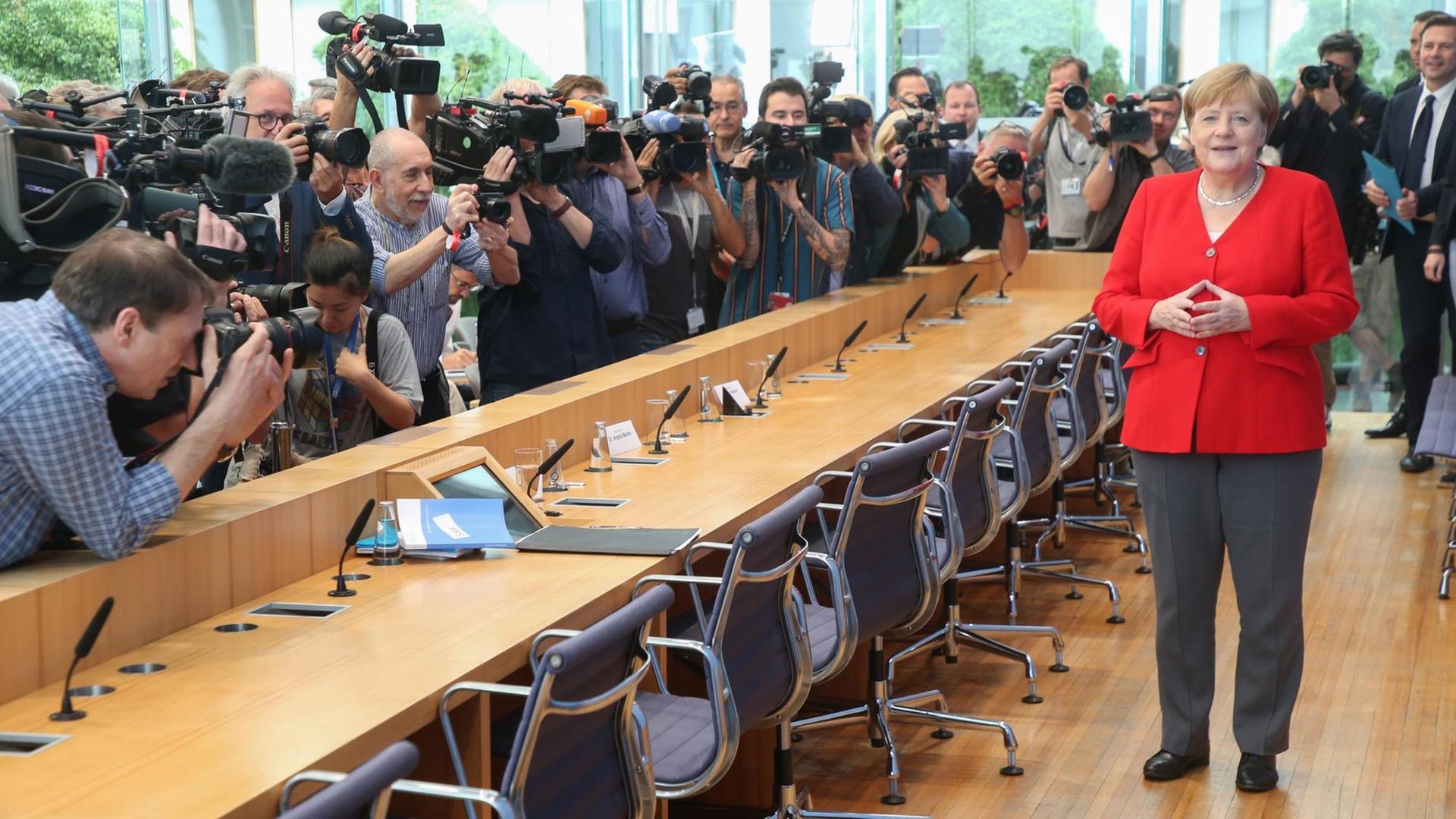 Foto- und Fernsehjournalisten drängen sich vor dem Rednerpult, Merkel steht dahinter und legt die Hände zur Raute zusammen.