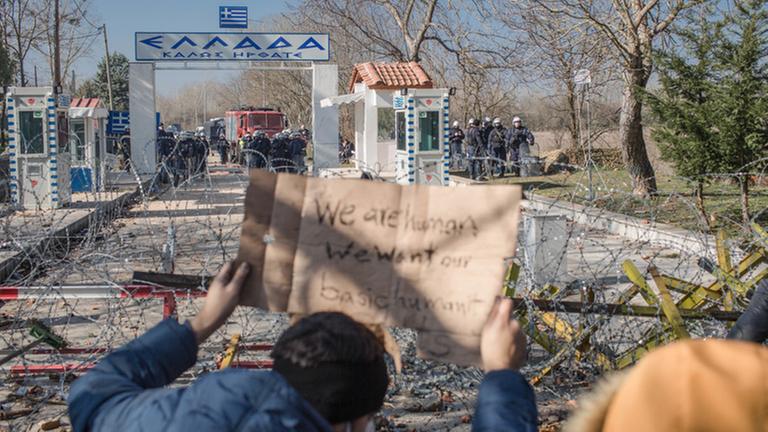 01.03.2020, Türkei, Pazarkule: Migranten stehen am bereits geschlossenen türkisch-griechischen Grenzübergang und halten ein Pappschild mit der Aufschrift "We are human we want our basic humanity" (Wir sind auch nur Menschen und wollen Menschlichkeit). An dem türkisch-griechischen Grenzübergang sind tausende weitere Flüchtlinge mit dem Ziel EU eingetroffen, nachdem bereits Tausende dort bereits die Nacht unter freiem Himmel verbracht haben. Foto: Ahmed Deeb/dpa | Verwendung weltweit