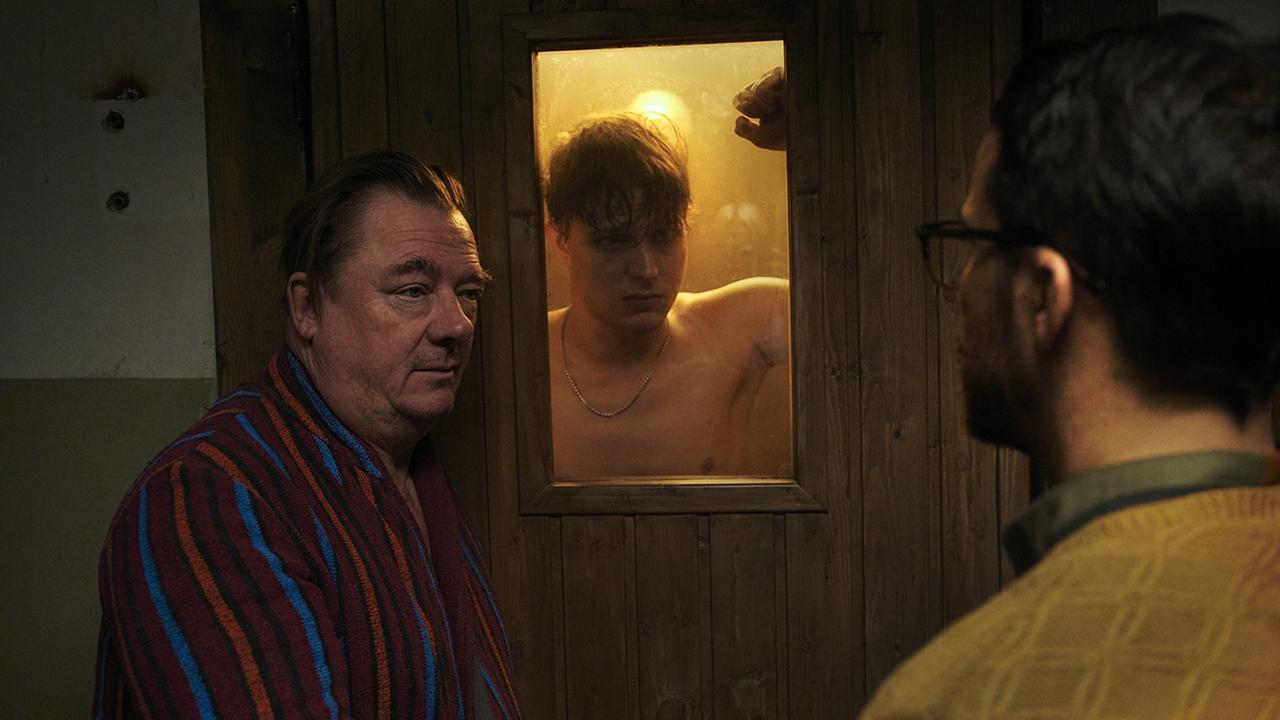 Szene aus "Wir können nicht anders" mit Peter Kurth (l), Merlin Rose und Kostja Ullmann. Zwei Männer stehen vor einer verschlossenen Sauna, wo sich ein anderer Mann befindet.
