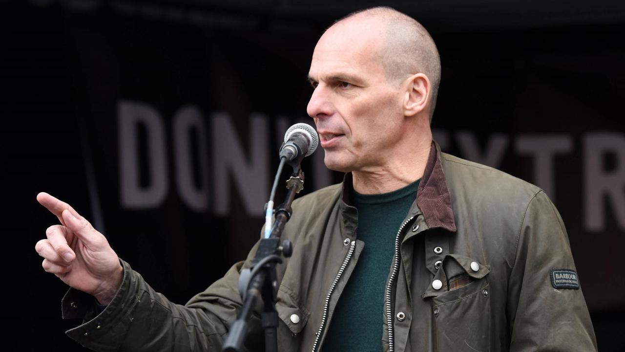 Janis Varoufakis, mit kahlgeschorenem Kopf und brauner Jacke spricht in ein Mikrofon und gestikuliert dabei mit der rechten Hand.