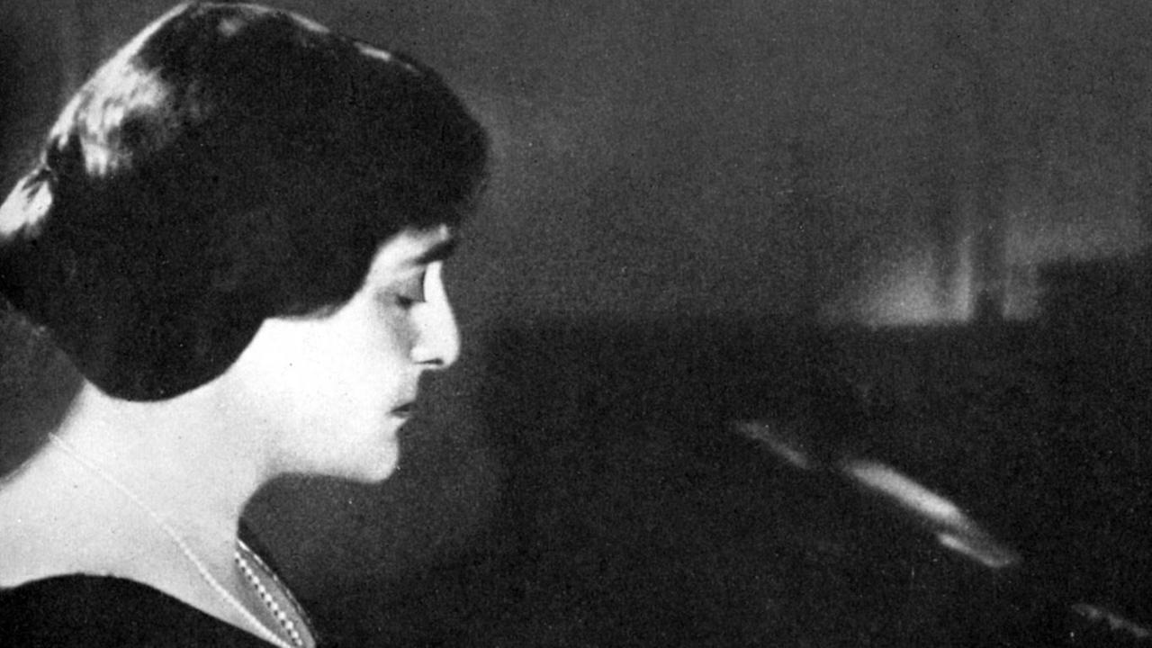 Schwarz-Weiß Fotografie der Pianistin im Halbprofil, sie trägt ein dunkles Kleid und eine Perlenkette und hat mitellange dunkle Haare. Sie hält die Augen halb geschlossen.