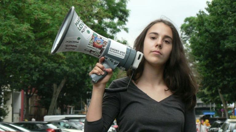 Hannah Pirot. Eine junge Frau mit langen braunen Haaren und einem Megafon in der Hand steht auf einer Straße.