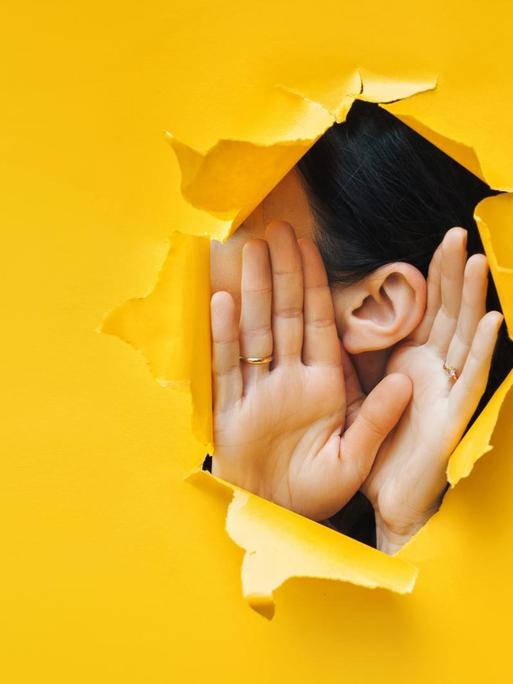 In eine gelbe Pappe ist ein Loch gerissen, durch das man ein Ohr und zwei Hände, die zum Zuhören aufgestellt sind, erkennt.