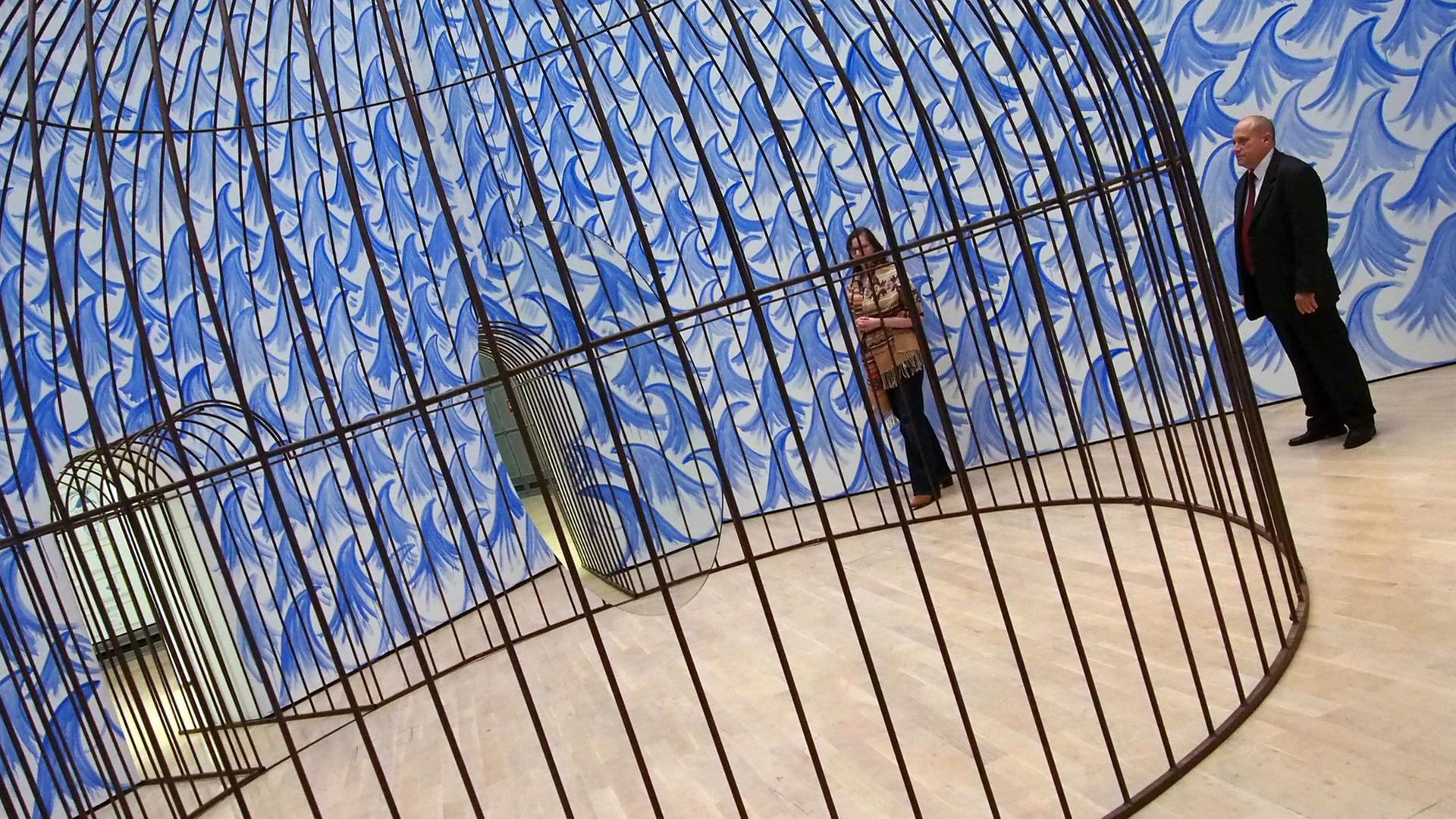 Eine Besucherin und ein Museumsmitarbeiter stehen am 11.11.2015 in der Kunstausstellung "Jeppe Hein. This Way" im Kunstmuseum Wolfsburg vor einem riesigen Käfig, in dem ein Spiegel kreist.