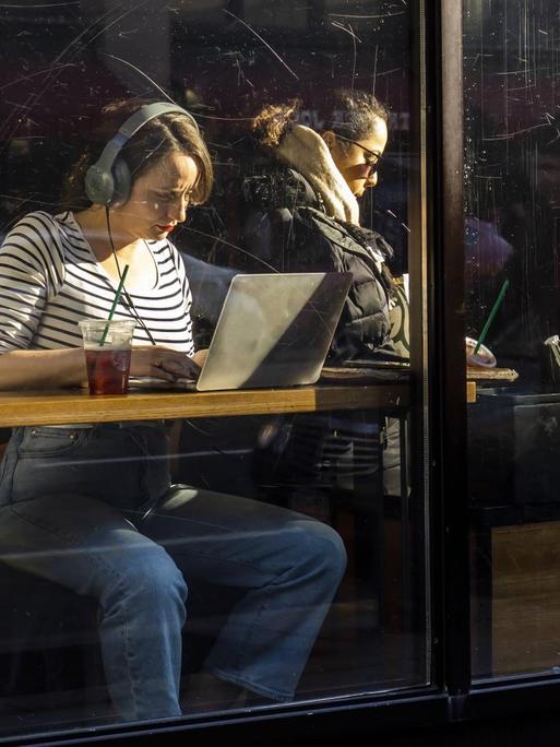 Eine Frau sitzt mit Laptop und Kopfhörern am Fenster eines Cafés.