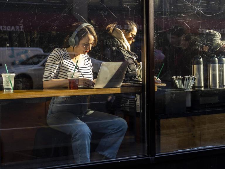 Eine Frau sitzt mit Laptop und Kopfhörern am Fenster eines Cafés.