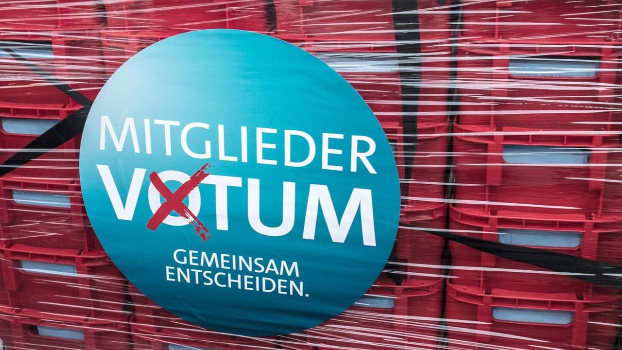 Das Bild zeigt in Plastik verpackte gestapelte rote Kisten mit den Wahlbriefen. Auf dem Plastik das Logo des Mitgliedervotums.