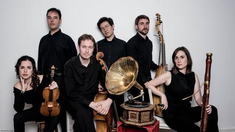 Das Ensemble Radio Antiqua variiert seine Besetzung mit verschiedenen historischen Blas-, Streich- und Zupfinstrumenten, hier auf dem Bild sind die sechs Musikerinnen und Musiker in schwarzer Kleidung zu sehen, teils sitzend, teils stehen um ein altes Grammophon