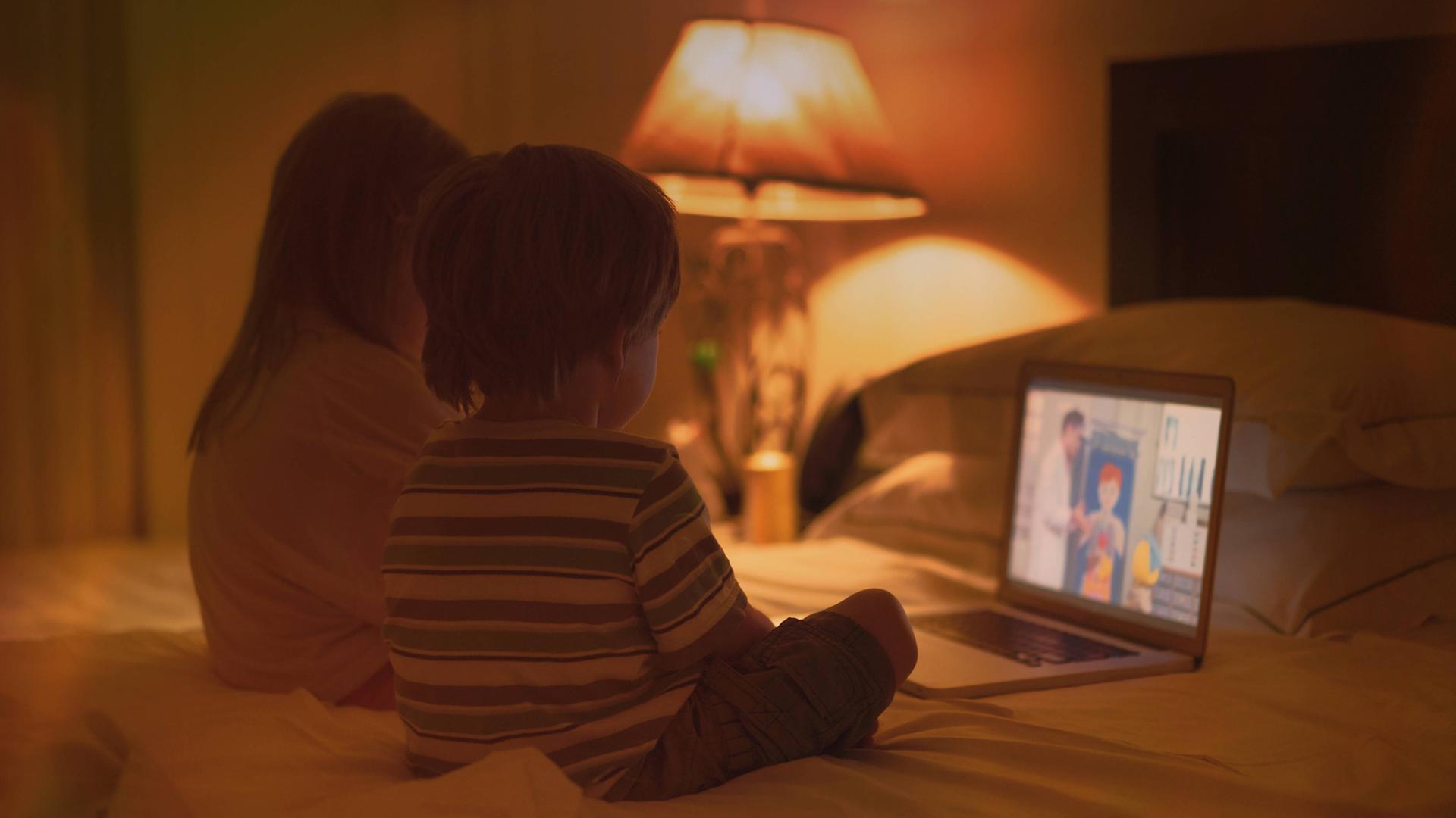Ein Mädchen und ein Junge sitzen auf einem Doppelbett und gucken einen Zeichentrickfilm auf einem Laptop.