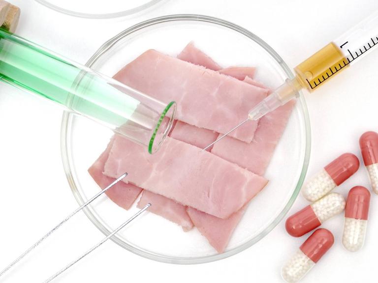 Symbolfoto für Fleisch aus dem Labor - eine Petrischale, ein Reagenzglas und Pillen liegen neben mehreren Scheiben Schinken