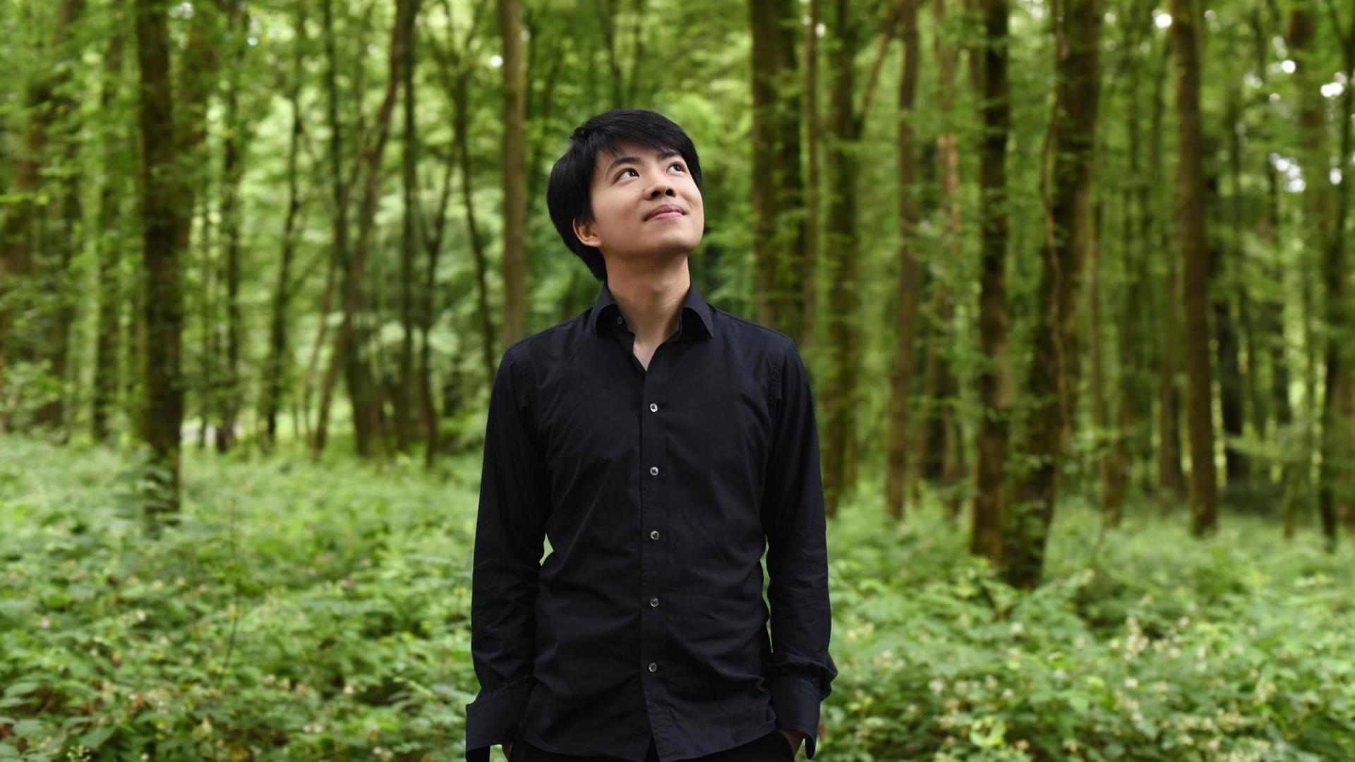 Ein junger Mann mit schwarzen Haaren und in schwarzem Hemd steht in einem lichten Wald und schaut entspannt Richtung Himmel.