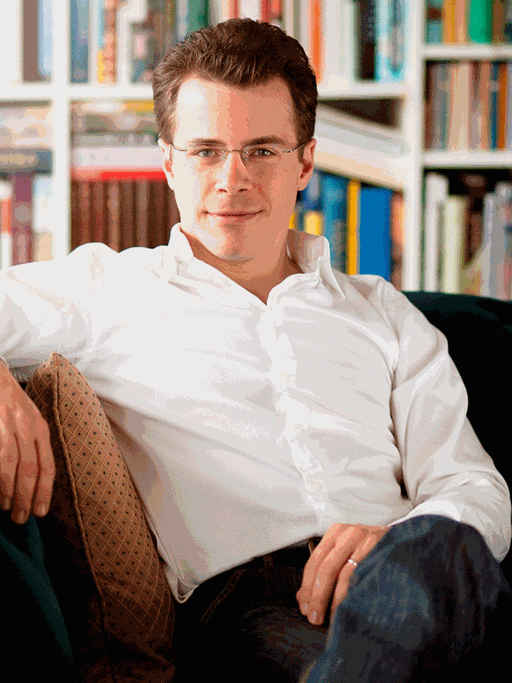 Der Dirigent Jakub Hrůša sitzt auf einem grünen Sofa umgeben von Regalen voller Noten und Bücher.