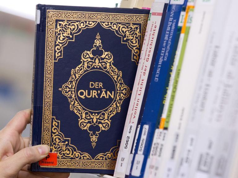 Eine Hand greift in einem Bücherregal einer Bibliothek nach einem Koran.