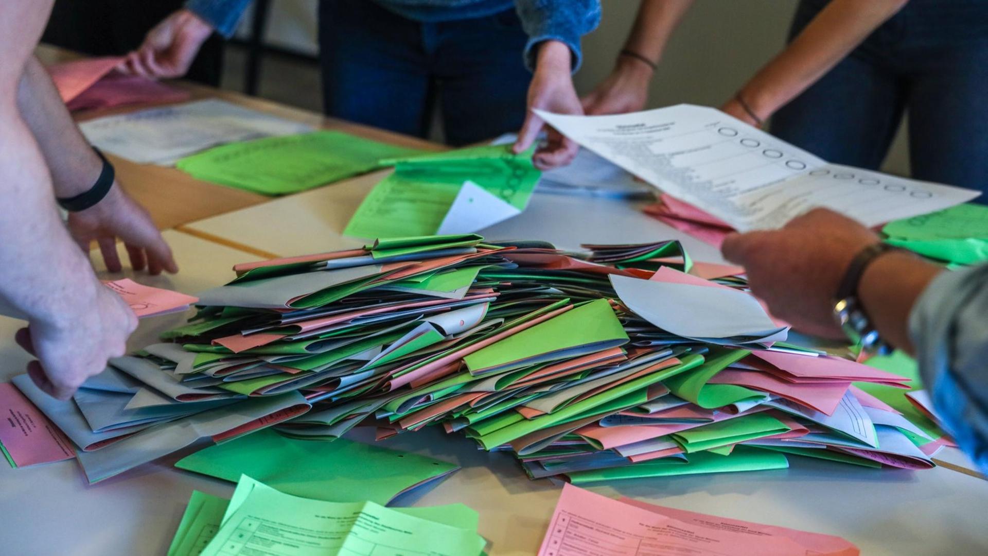 Viele Wahlzettel liegen in Stapeln auf einem Tisch, vier Menschen sortieren sie.