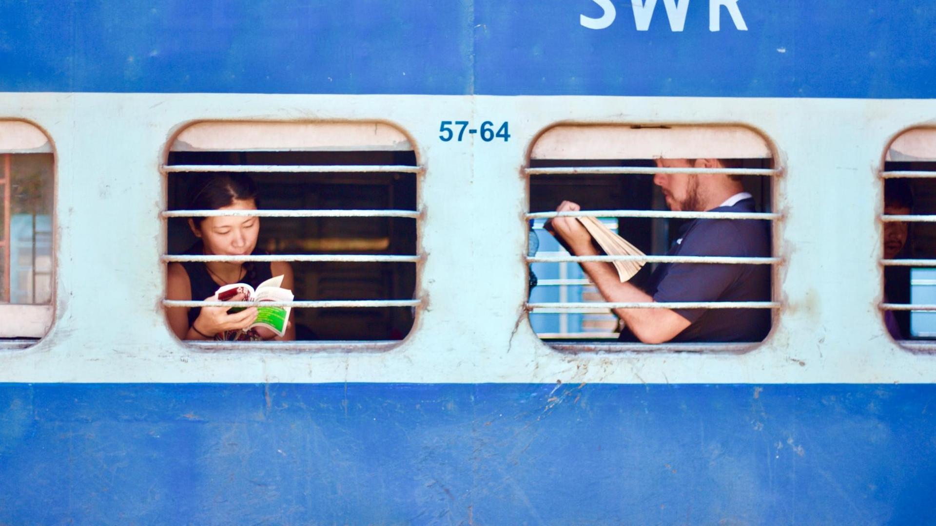 Ein blauer Zug in Indien in dem Menschen am Fenster sitzend lesen.