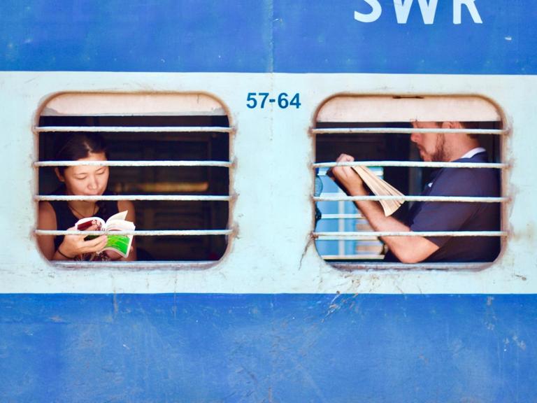 Ein blauer Zug in Indien in dem Menschen am Fenster sitzend lesen.