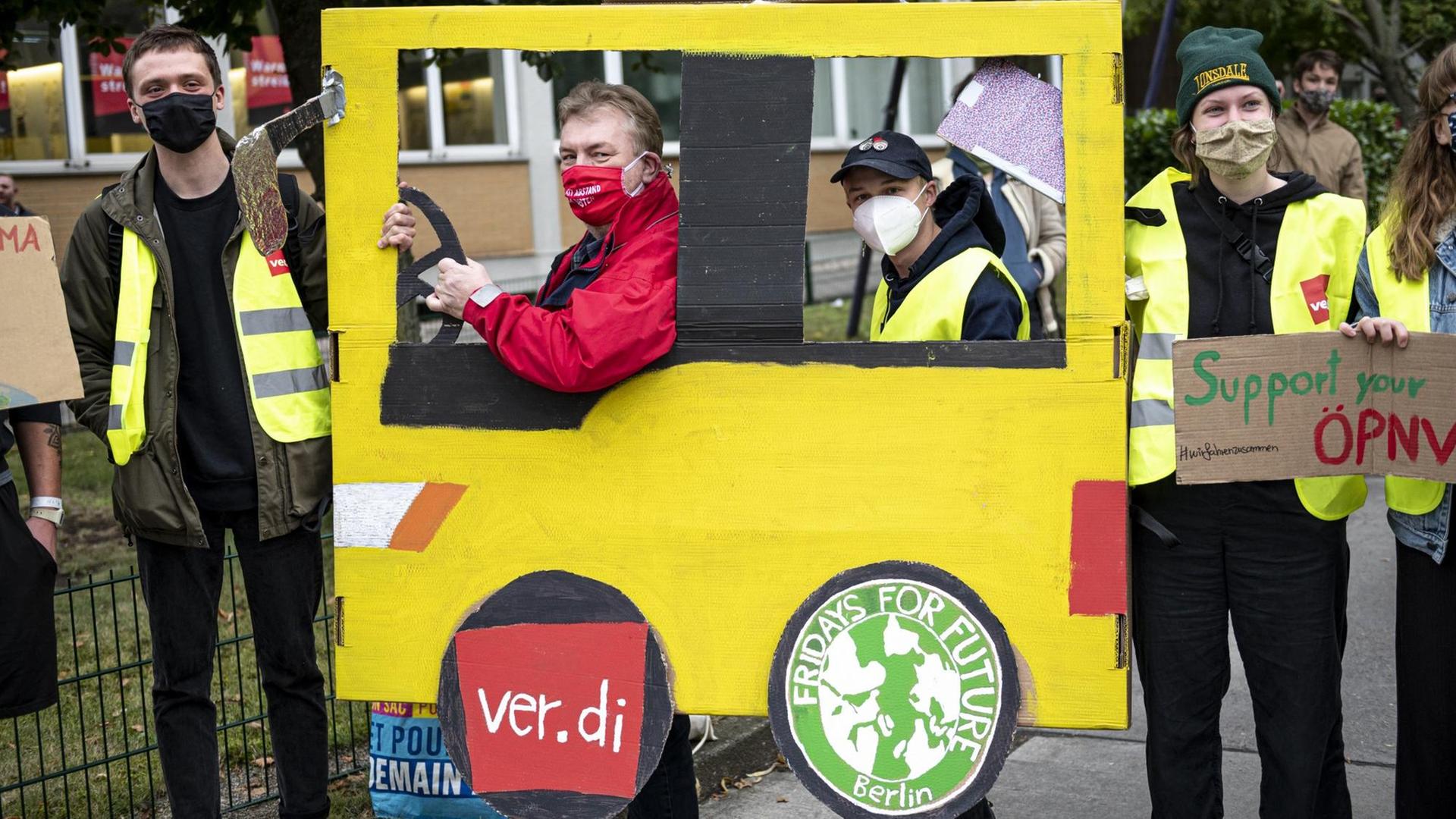 Bus-Fahrerinnen und Bus-Fahrer demonstrieren mit einem großen Bus aus gelber Pappe. 