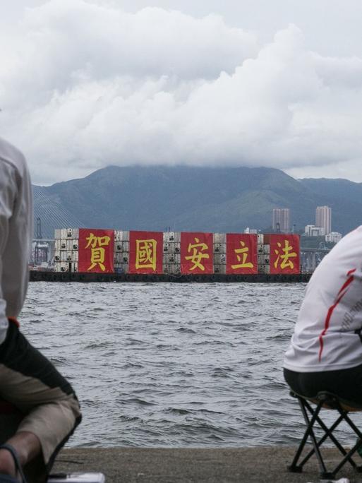 Angler in Hongkong betrachten ein vorüber fahrendes Schiff, welches auf roten Transparenten das von China erlassene neue Sicherheitsgesetz als nationalen Erfolg feiert.