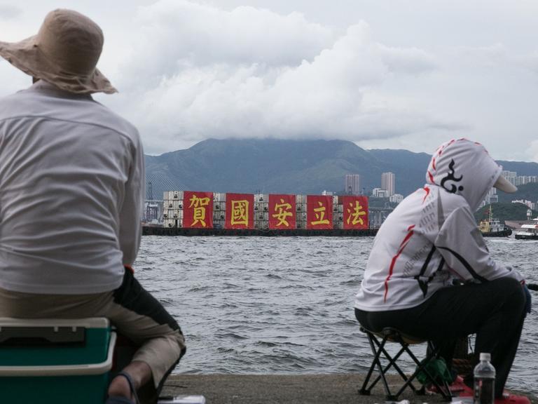 Angler in Hongkong betrachten ein vorüber fahrendes Schiff, welches auf roten Transparenten das von China erlassene neue Sicherheitsgesetz als nationalen Erfolg feiert.