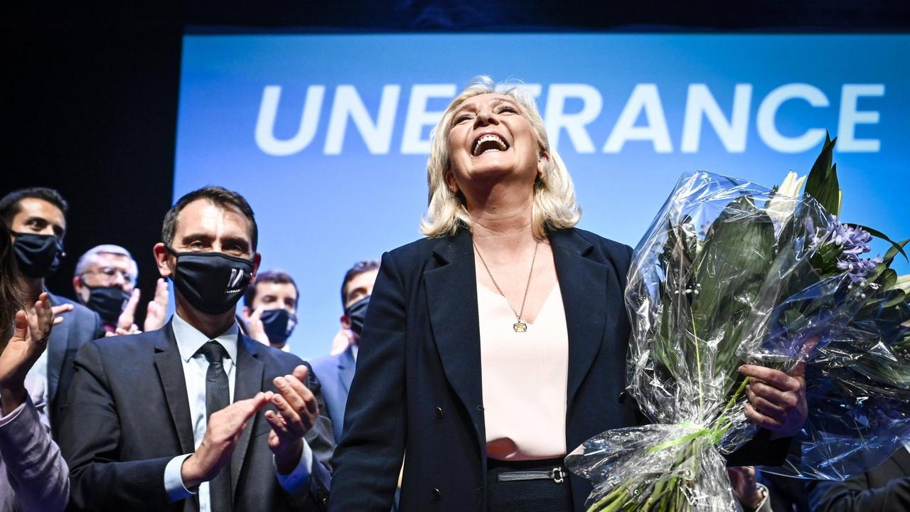 Die Vorsitzende des Rassemblement National, Marine Le Pen, steht auf einer Bühne vor einem Transparent "Une France". Sie hält einen Blumenstrauß und lacht, hinter ihr stehen viele klatschende Menschen, die FFP-Masken tragen. 04/07/2021