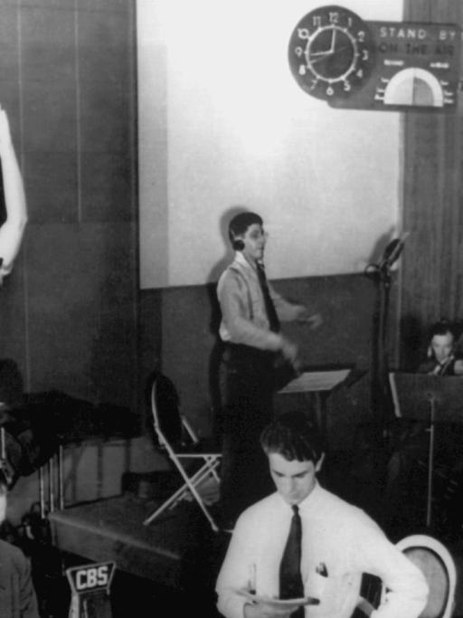 Schwarz-weiß-Aufnahme einer Hörspiel-Übertragung in einem Studio. Sprecher an Mikrofonen und Musiker mit Geigen.