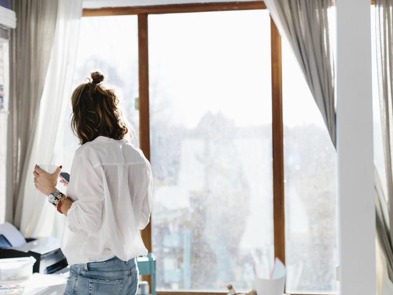 Eine Frau steht mit ihrem Smartphone und einer Tasse Kaffee in der Hand in ihrem Home Office.