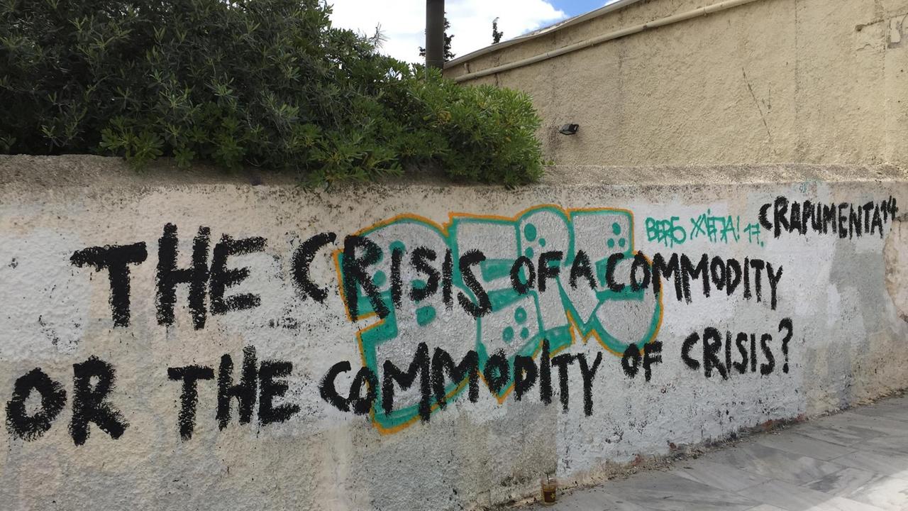"The crisis of a commodity or the commodity of the crisis" und "crapumenta 14" steht mit schwarzer Farbe auf einer Mauer in Athen geschrieben.