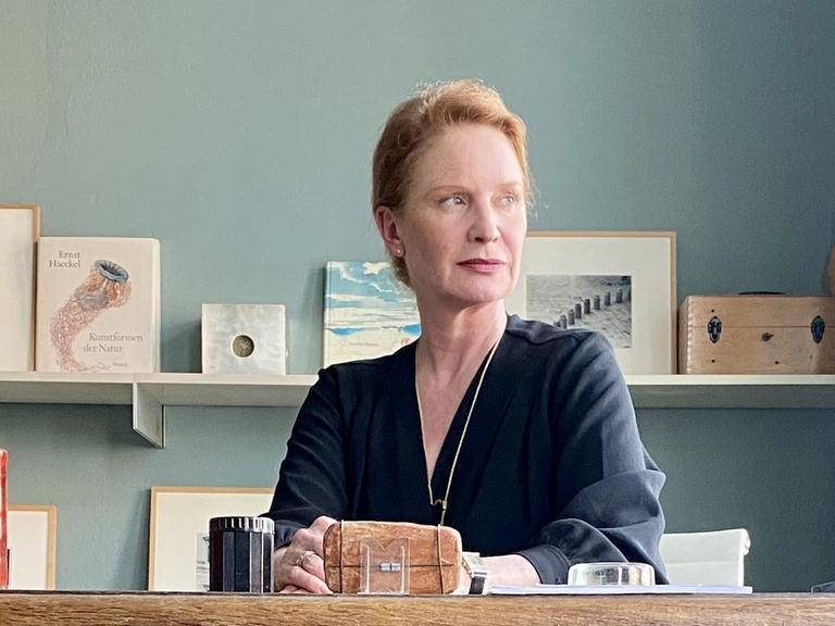 Die Schriftstellerin Eva Sichelschmidt sitzt an ihrem alten hölzernen Schreibtisch und blickt Richtung Fenster.