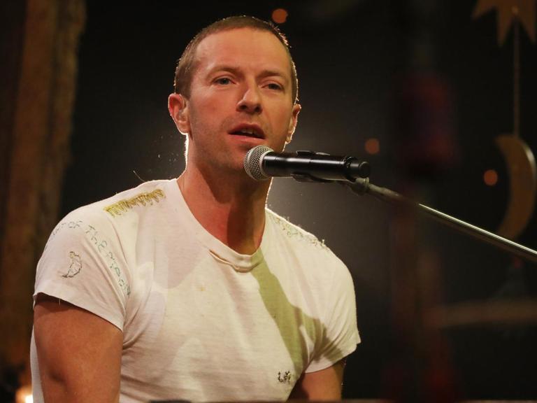 Coldplay-Sänger Christ Martin sitzt bei einem TV-Auftritt in der Graham Norton Show am Flügel. Er trägt ein weißes T-Shirt.