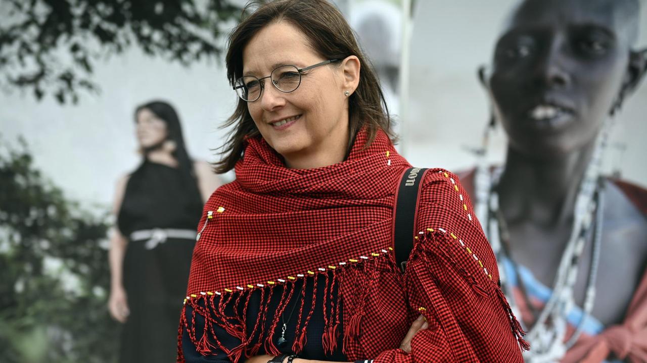 Eine Frau mit Brille und rotem Schal steht die Arme verschrängt vor großen Fotografien. 