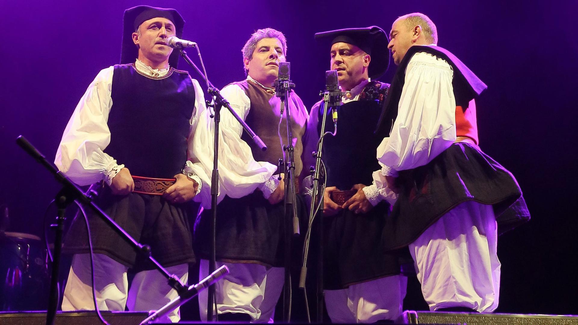 Das sardische Vokalquartett Tenores di Bitti "Mialinu Pira" steht in traditioneller Kleidung auf einer Bühne.