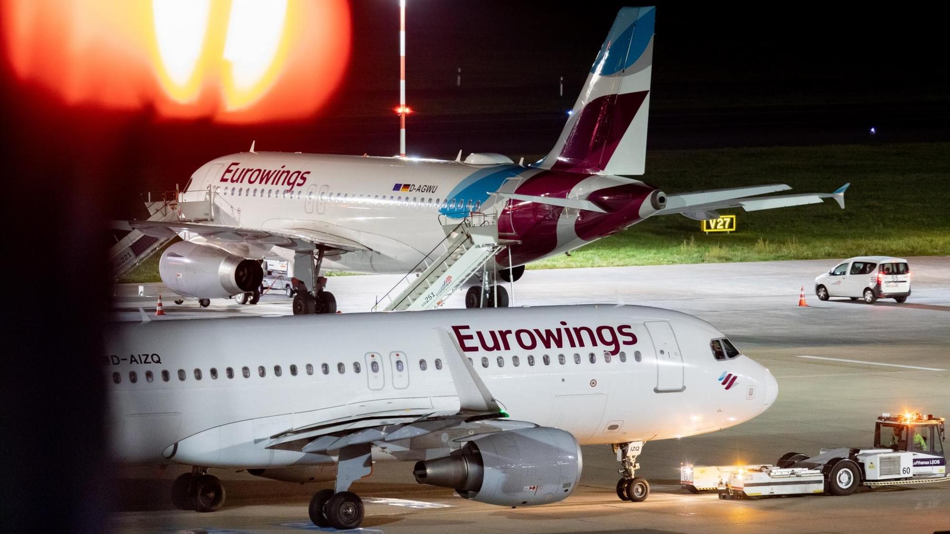Zwei Maschinen der Fluggesellschaft Eurowings stehen auf dem Vorfeld, im Vordergrund links oben ist die Beleuchtung eines Gebäudes zu sehen.