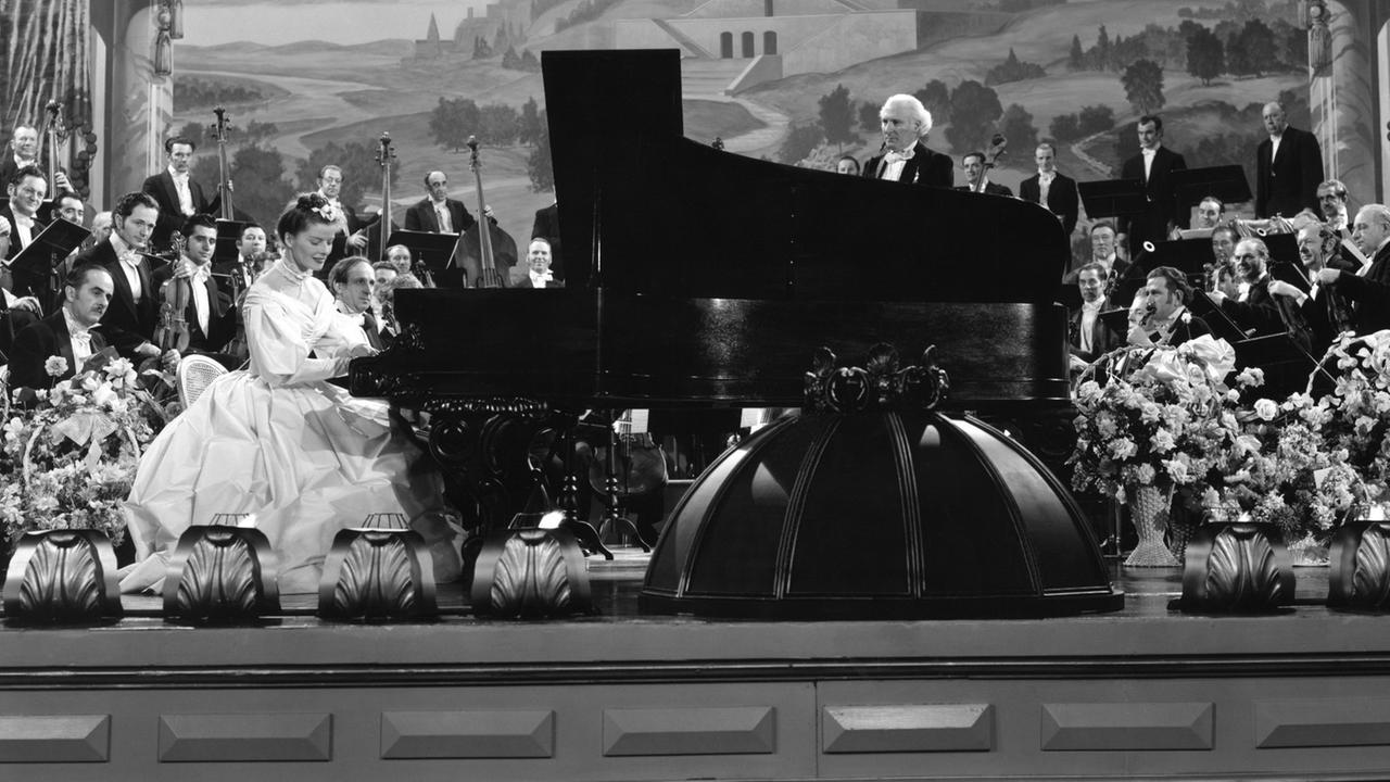Ein historisches Foto zeigt eine festlich gekleidete Frau im Stil des 19. Jahrhunderts auf einer Bühne Klavier spielend, von einem Orchester begleitet, wobei der Dirigent über den Flügel hinweg zur Musikerin schaut.