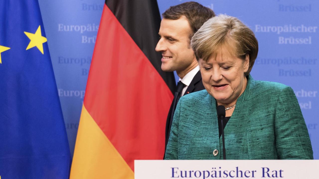 Merkel steht lächelnd am Rednerpult, hinter ihr läuft ebenfalls lächelnd Macron vorbei. An der Wand eine deutsche und eine EU-Flagge.