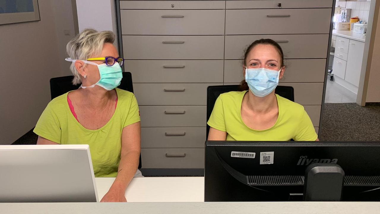 Eine Sprechstundenhilfe und eine Arzthelferin der Praxis Heinert im badischen Bahlingen sitzen hinter dem Anmeldungstresen. Sie tragen Mundschutzmasken.
