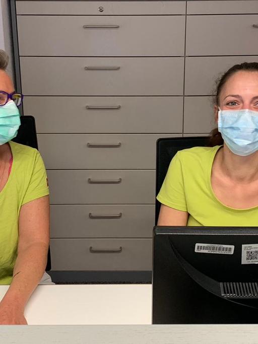 Eine Sprechstundenhilfe und eine Arzthelferin der Praxis Heinert im badischen Bahlingen sitzen hinter dem Anmeldungstresen. Sie tragen Mundschutzmasken.