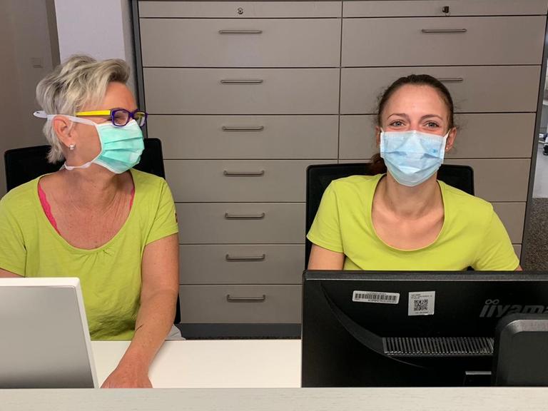 Eine Sprechstundenhilfe und eine Arzthelferin der Praxis Heinert im badischen Bahlingen sitzen hinter dem Anmeldungstresen. Sie tragen Mundschutzmasken.