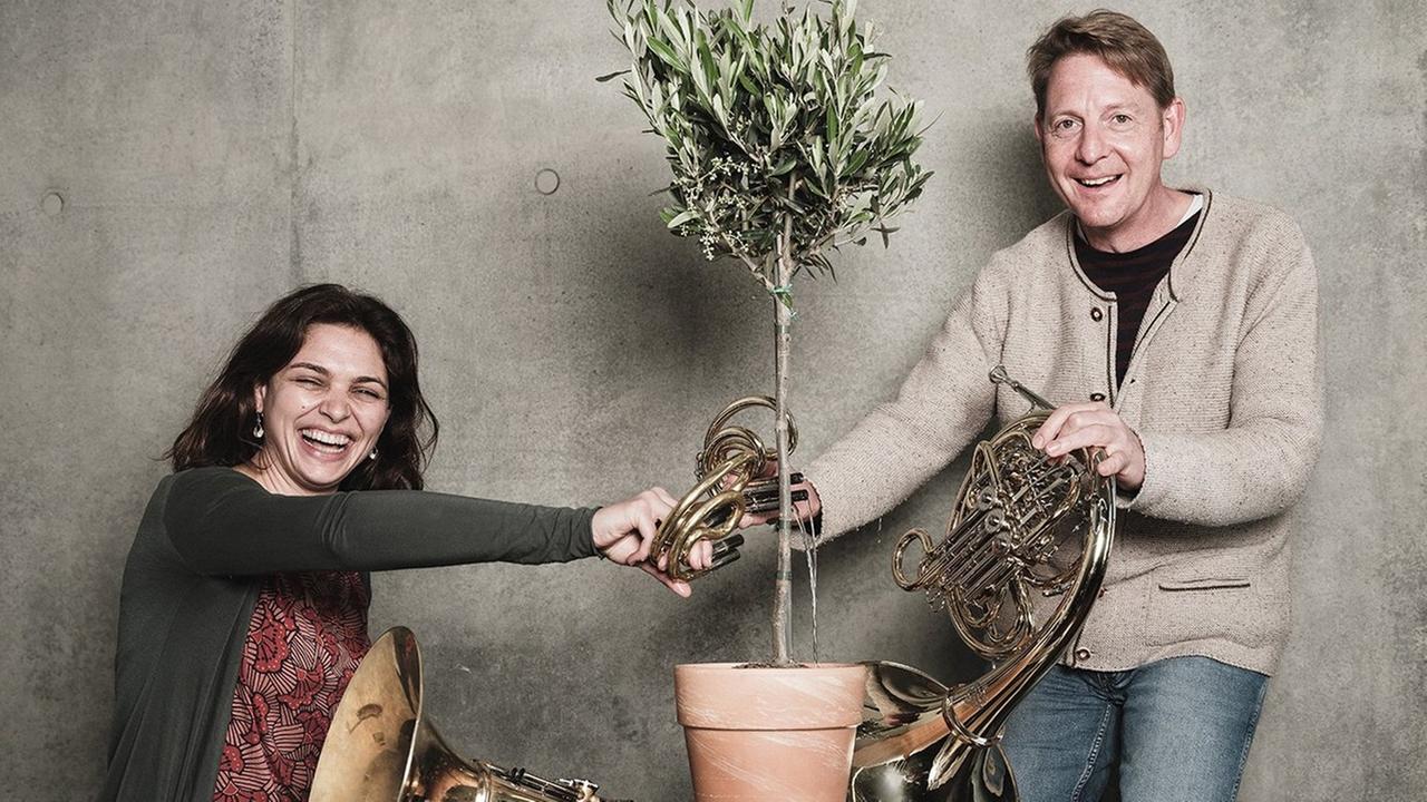 Eine Frau und ein Mann lachen in die Kamera. Zwischen ihnen steht ein Olivenbaum, den sie mit einem kleinen Horn gießen. Beide haben jeweils ein großes Horn in der Hand.