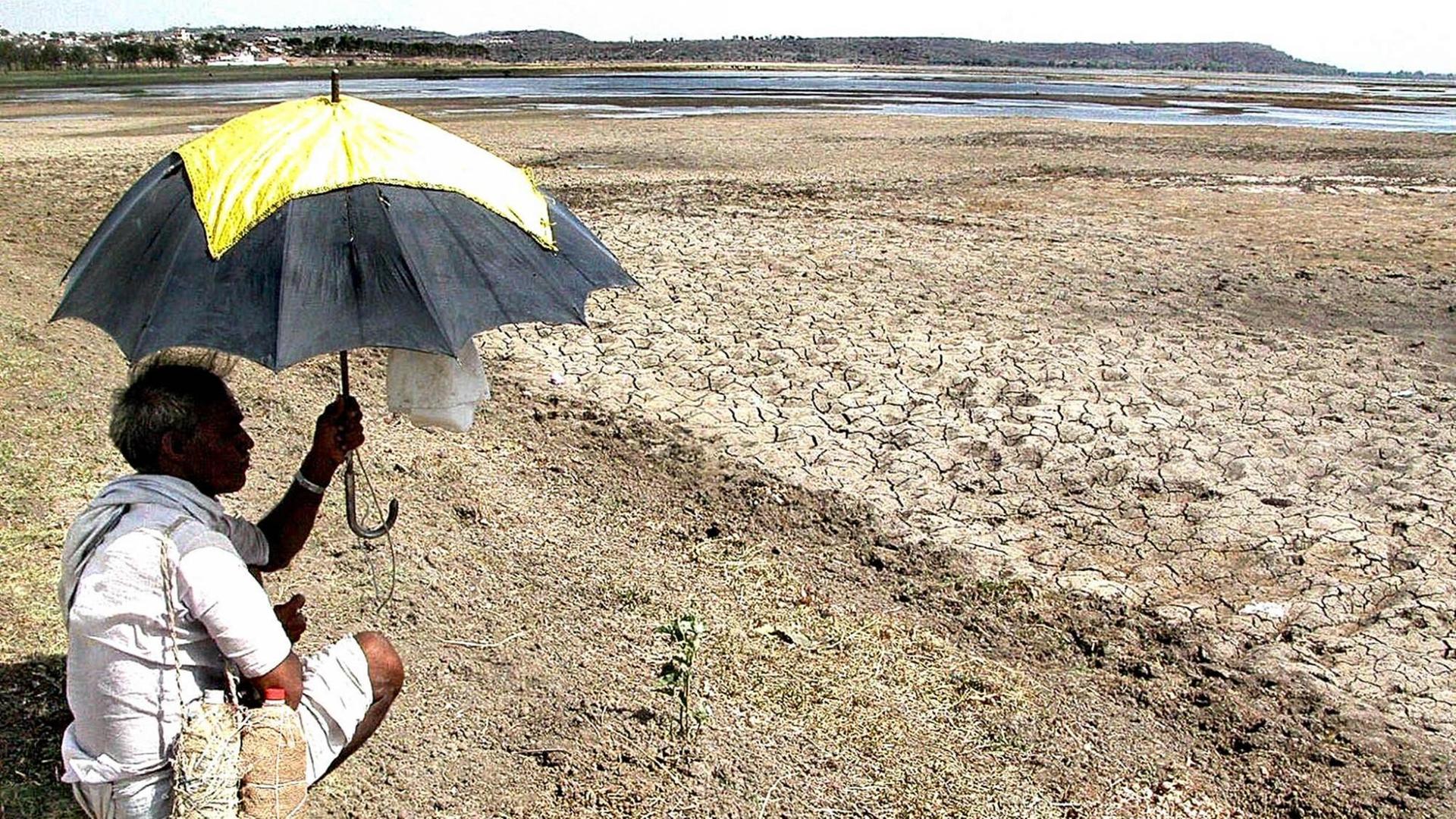 Ein Mann sitzt mit einem Sonnenschirm in der Hand am Ufer eines fast ausgetrockneten Sees im indischen Bhopal
