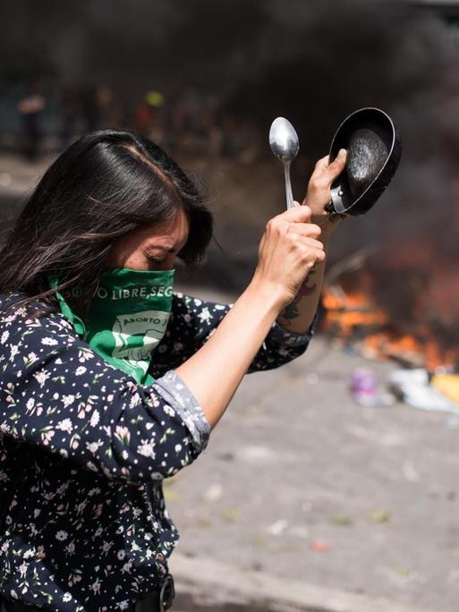 Proteste in Chile gegen Preiserhöhungen im öffentlichen Nahverkehr. Eine Frau schlägt auf ein Gefäß, im Hintergrund brennender Müll auf der Straße.