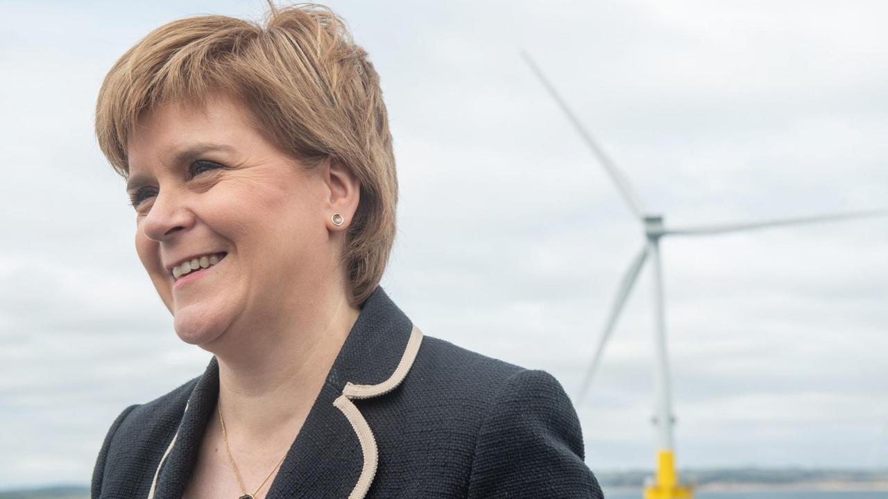Schottlands Erste Ministerin Nicola Sturgeon vor blauem Himmel, im Hintergrund ist ein Windrad in der Nordsee zu sehen.