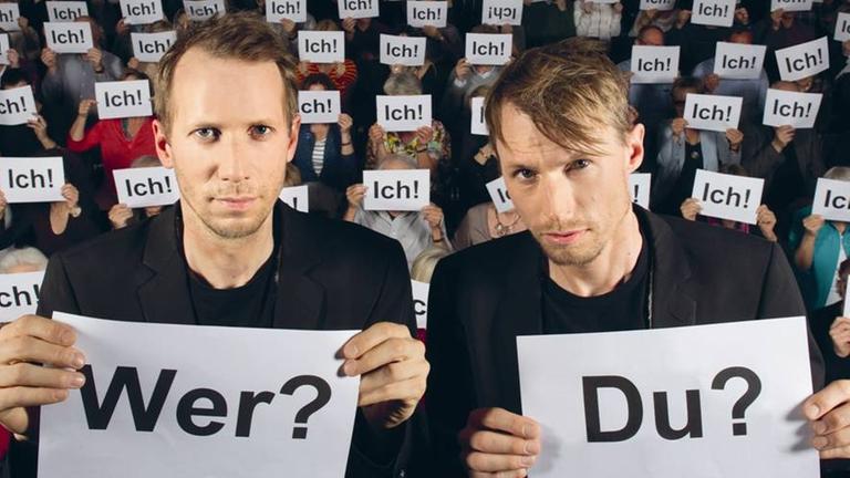 Das Kabarett-Duo "Ohne Rolf": Jonas Anderhub und Christof Wolfisberg. Sie stehen nebeneinander und halten jeweils ein Blatt Papier hoch. Auf dem einen steht "Wer?", auf dem anderen "Du?". Im Hintergrund sieht man eine Gruppe von Menschen, die sich jeweils ein Blatt Papier mit dem Aufdruck "Ich!" vor das Gesicht halten.