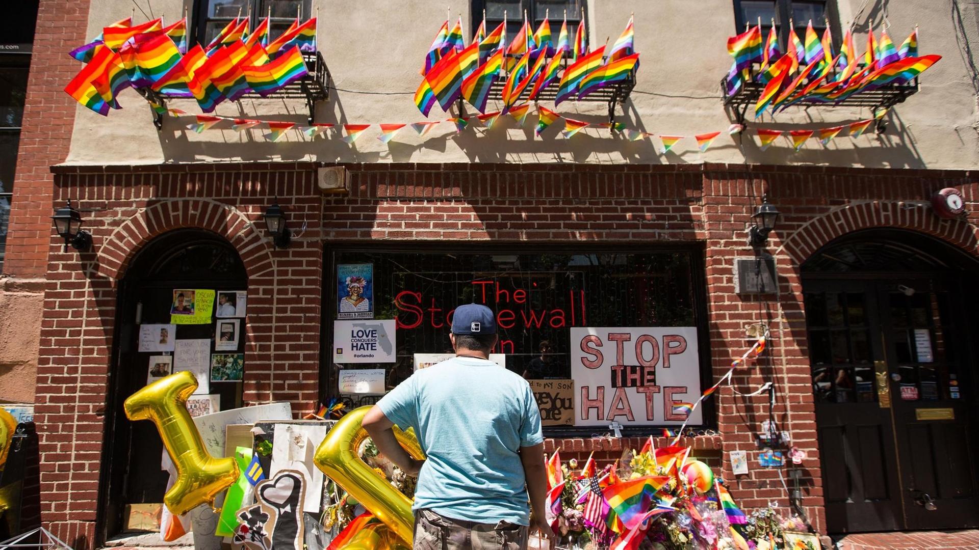 Ein Mann steht vor dem Stonewall Inn. Vor dem Gebäude liegen viele Gedenkobjekte. An der Fassade hängen Flaggen.