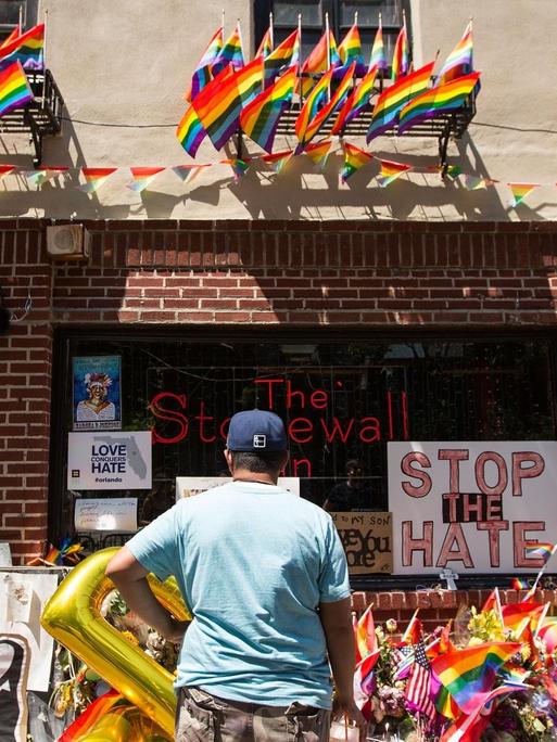 Ein Mann steht vor dem Stonewall Inn. Vor dem Gebäude liegen viele Gedenkobjekte. An der Fassade hängen Flaggen.