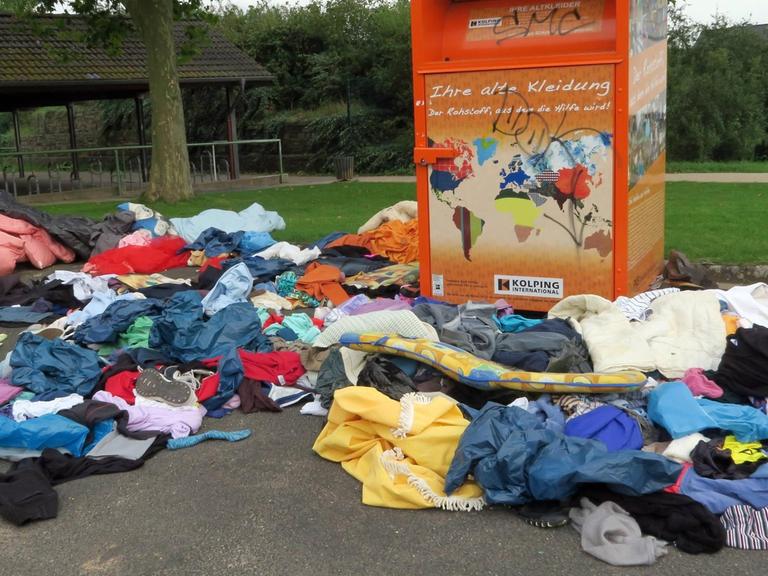 Textilien, Schuhe und Polster liegen neben einem Container für Altkleider auf der Straße.