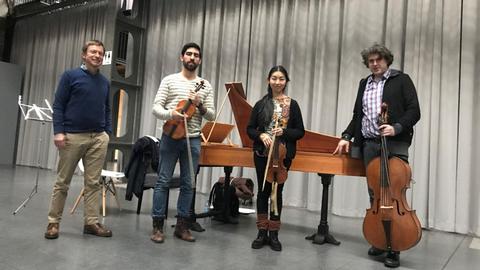 3 Musiker und eine Musikerin (in der Mitte) stehen vor einem Cembalo, dahinter ein ein heller Vorhang, drei haben ihre Streichinstrumente in der Hand, (2 Geigen, 1 Violoncello). Sie tragen Alltagskleidung