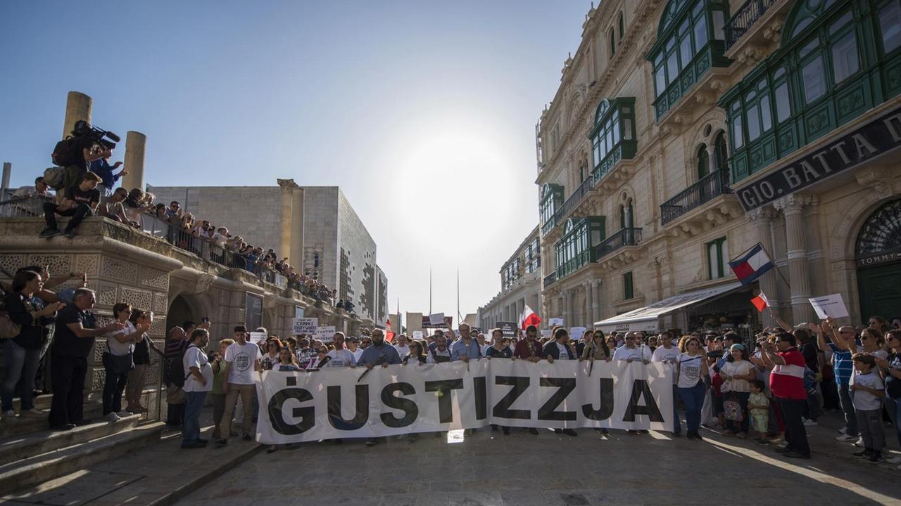 Die Menschen stehen quer über eine gepflasterte Straße hinter einem breiten Transparent mit der Aufschrift "Gustizzja". Auch an den Seiten stehen Demonstranten sowie Medienvertreter.